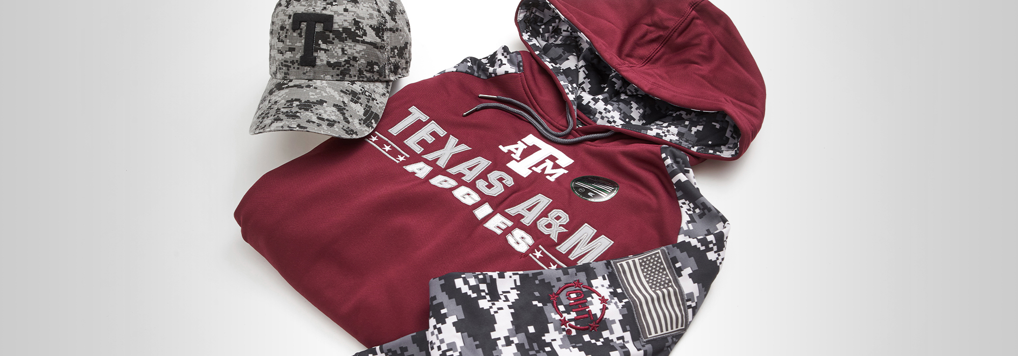 TAMU and Operation Hat Trick branded merchandise laid out on a white background. Items include a camo hat and maroon and camo sweatshirt. 