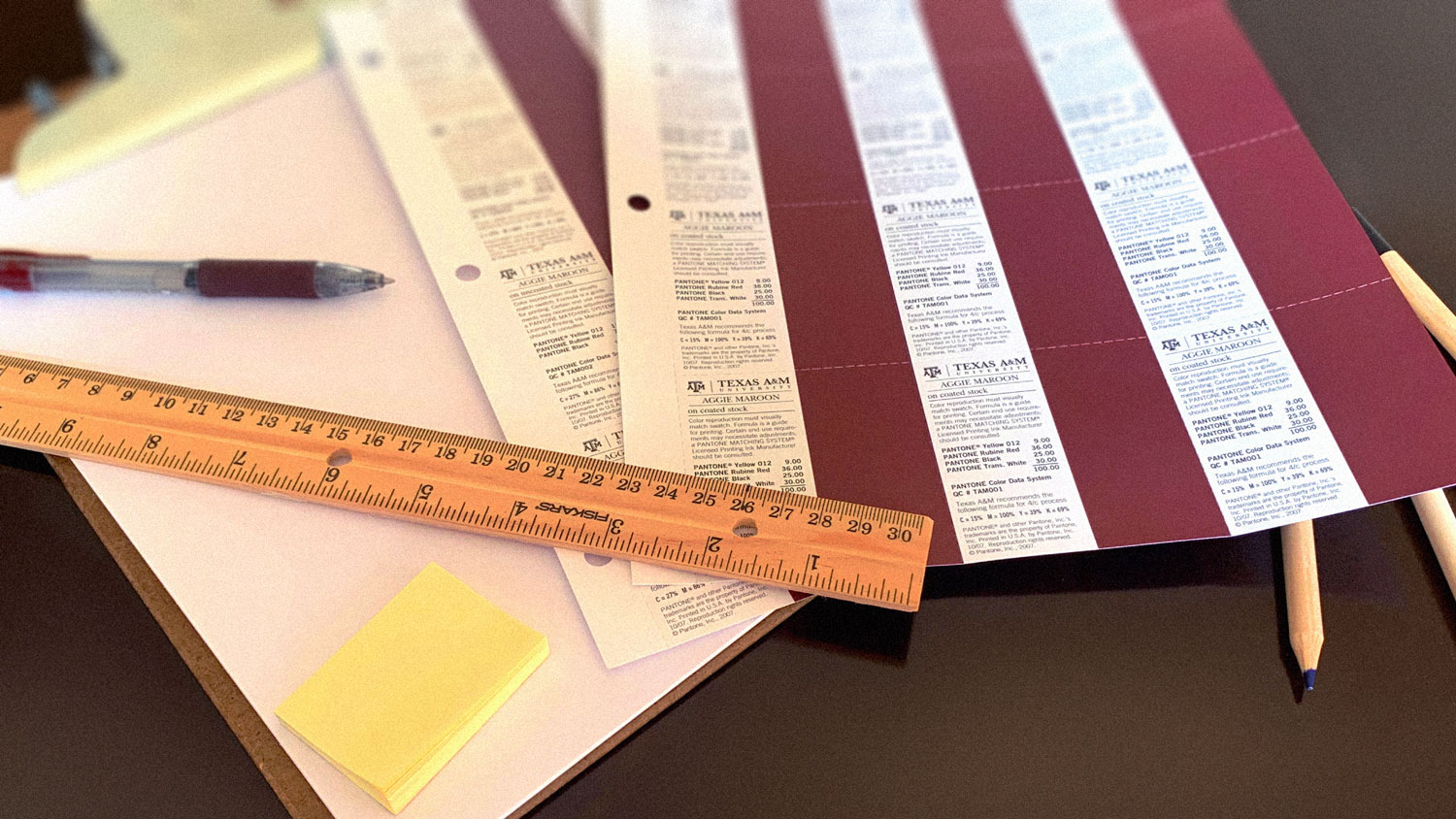 Texas A&M University maroon color swatches on a table next to maroon pens, a ruler and colored pencils