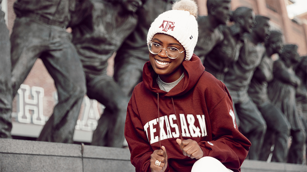 Texas A&M student stands in front of the Saw Em Off statue outside Kyle Field with a Texas A&M hoodie and beanie on